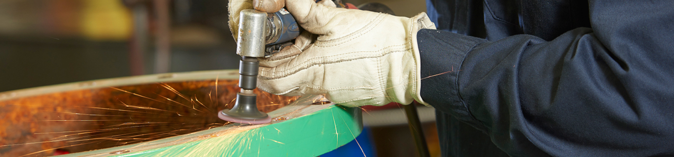 Image depicts a close-up shot of a worker's gloved hands using a grinding tool on a metal valve surface