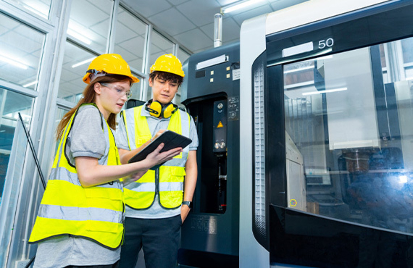 Two young employees wearing hard hats analyzing a computer in a factory, focusing on preventative maintenance services and uptime.