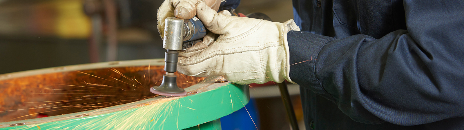 Image depicts a close-up shot of a worker's gloved hands using a grinding tool on a metal valve surface