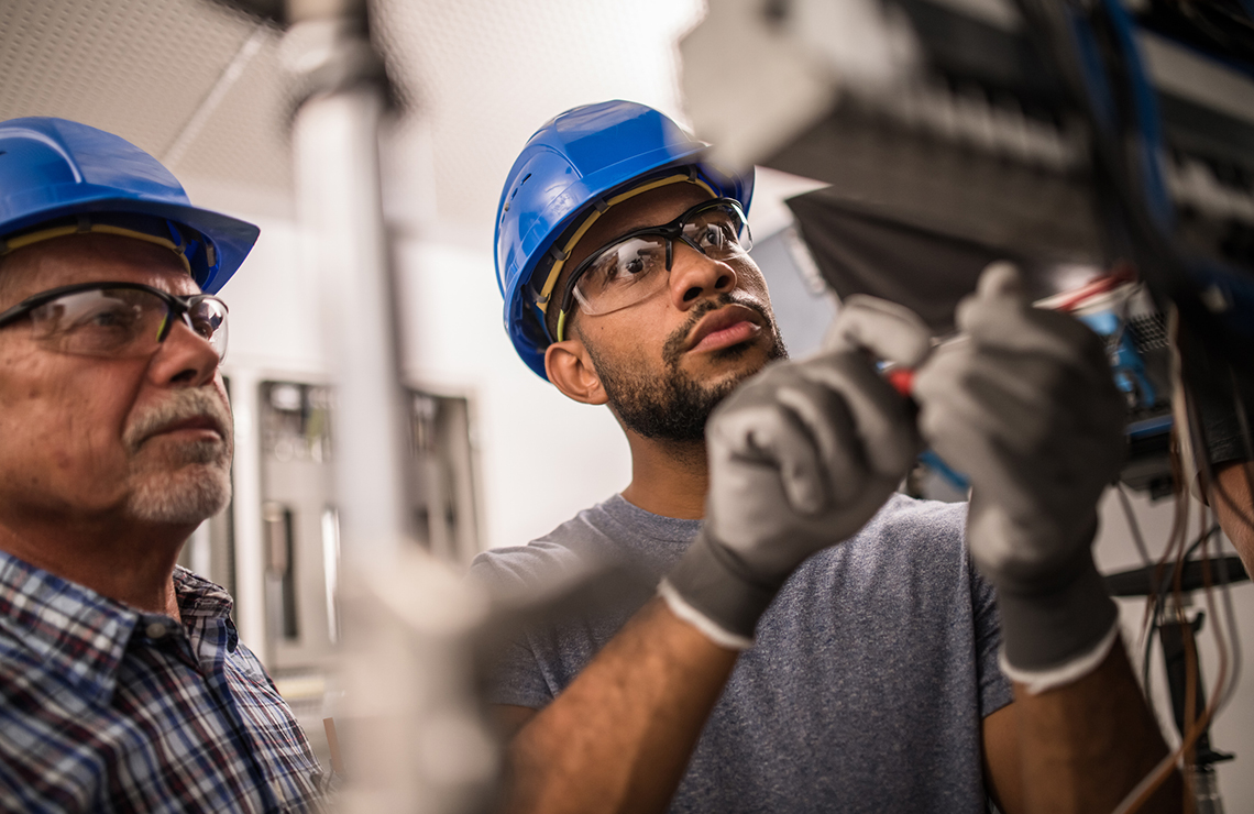 Image shows two industrial engineers in blue hard hats working on an electrical panel
