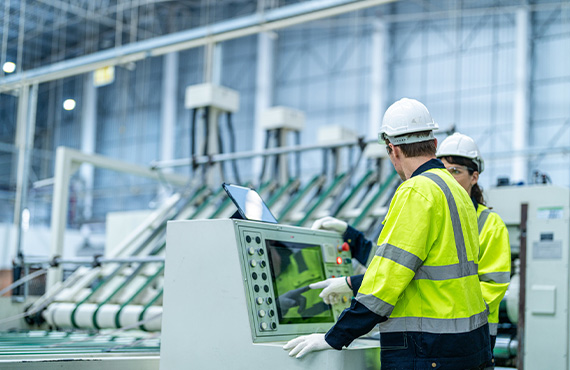 Two workers in safety gear operate a machine in a factory, focusing on PLC modernization solutions for plant managers.
